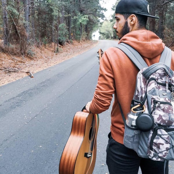 A bearded young YWAMer in the School of Worship walks along a wooded country road with a guitar in hand.  He is wearing an orange sweatshirt and a camo backpack.