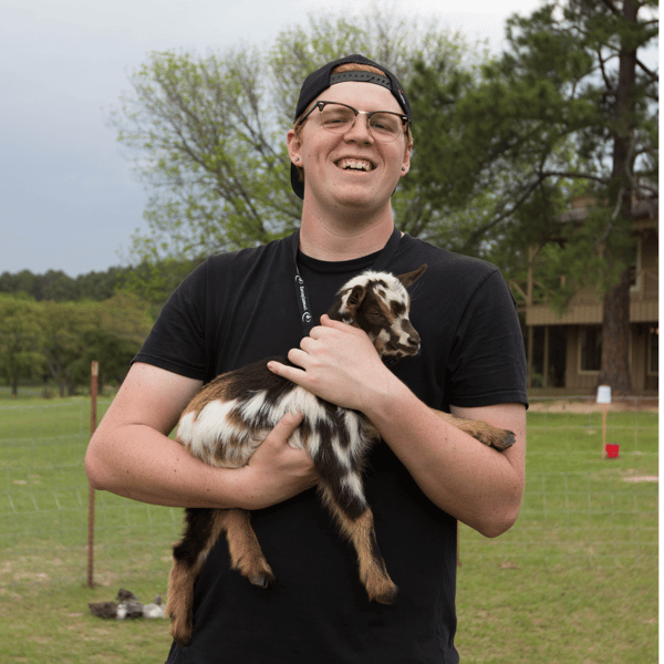 A YWAM student in the Agtech school is smiling and standing in a field while holding a baby goat.
