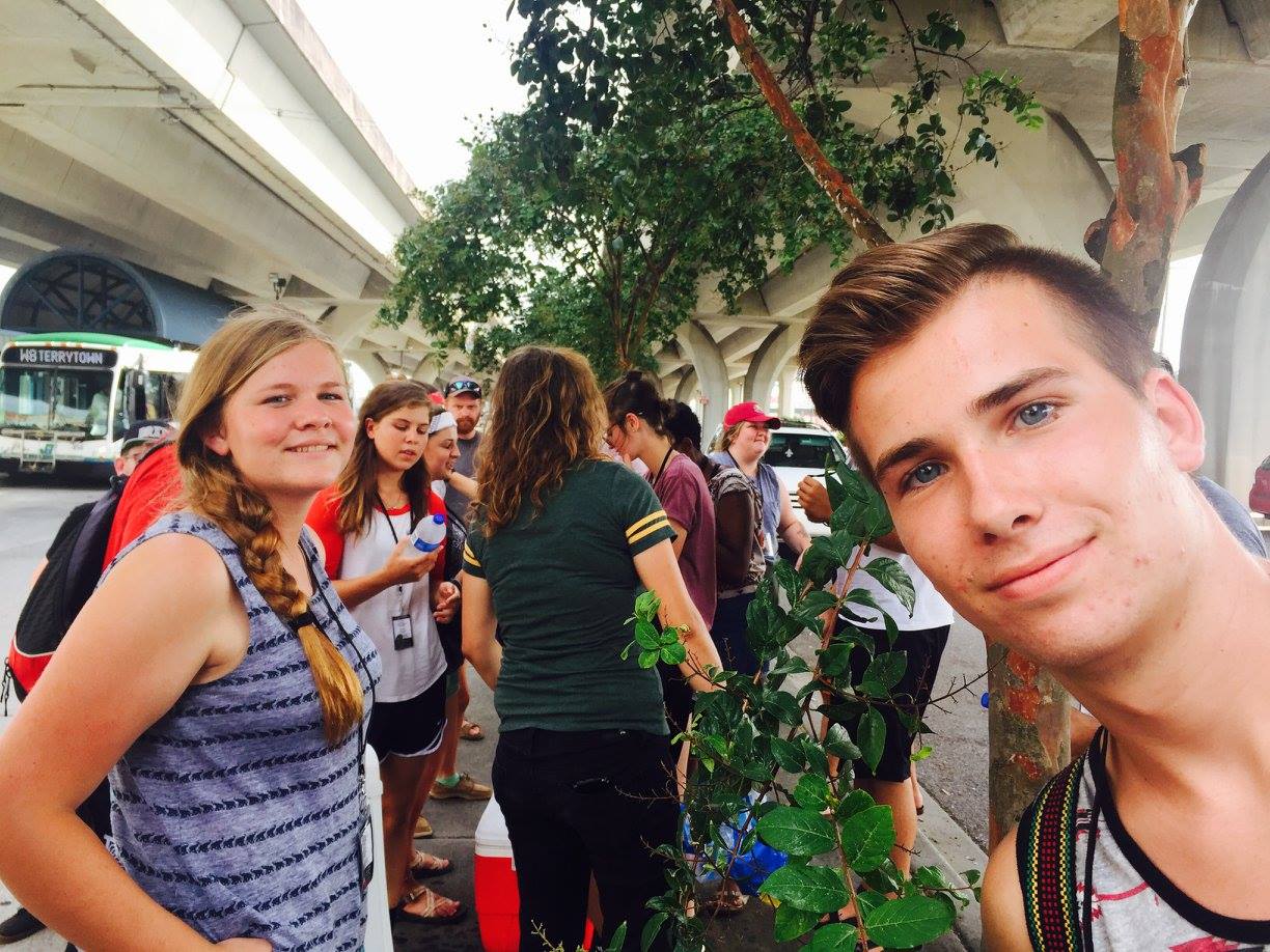 A older teenage boy and girl smiling at the camera, while the other SST students in the background are handing out or receiving water bottles.  They are all next to a small tree beneath a large freeway overpass.
