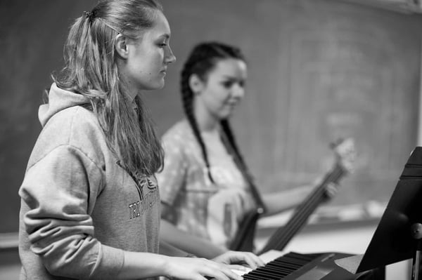 Two women in the school of worship are sitting and playing worship together.  One is playing piano, and the other is playing bass.