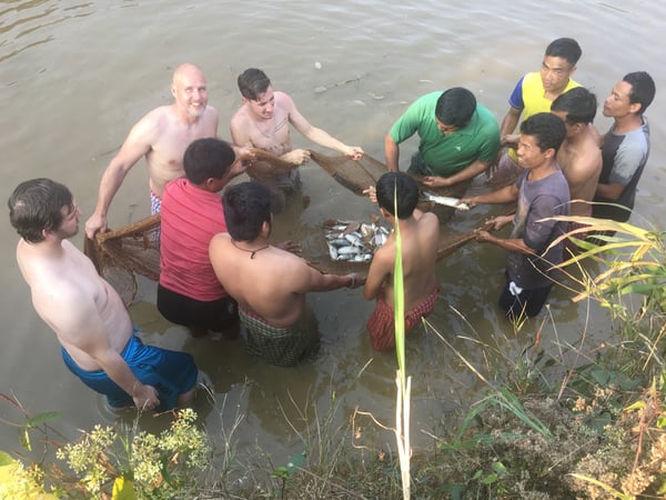 A group of young men and Agtech student and staff are all excitedly standing around in a lake.  They are all shirtless, up to their knees in water and hold the edges of a fishing net, filled with dozens of fish.