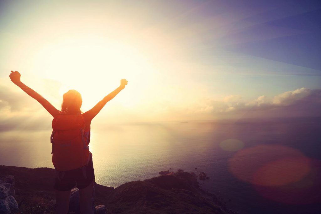 A youth wearing a backpack and hiking shorts standing on the edge of a cliff overlooking a cloud bank.  The sun is rising and their hands are stretch out into the air in victory.